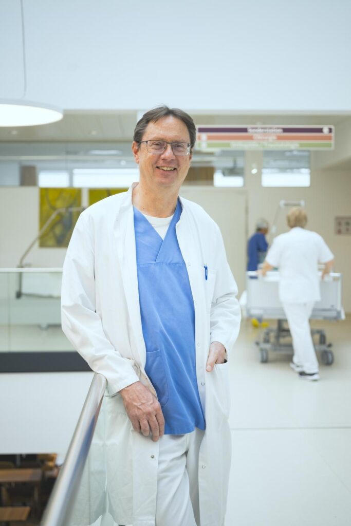 Prof. Dr. Ivo Graziadei steht in seiner Arbeitskleidung (weißer Kittel, blauer Kasak) im Gang des Krankenhauses Hall, im Hintergrund schieben Mitarbeiter Krankenbetten über den Flur
